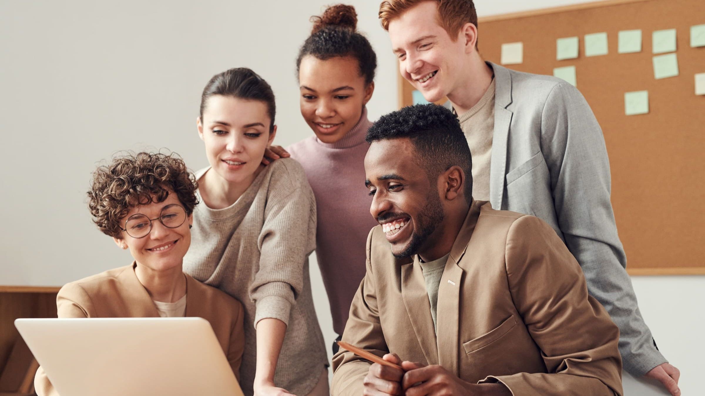 Five smiling young colleagues gather around a laptop in a bright office with a corkboard
