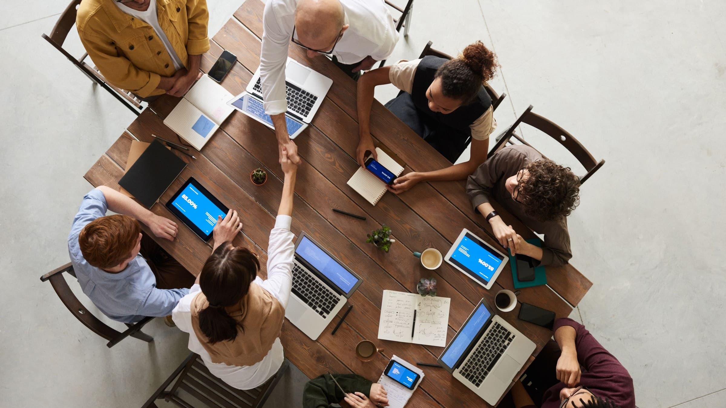 Overhead view of a team at a wooden table with laptops as two people shake hands