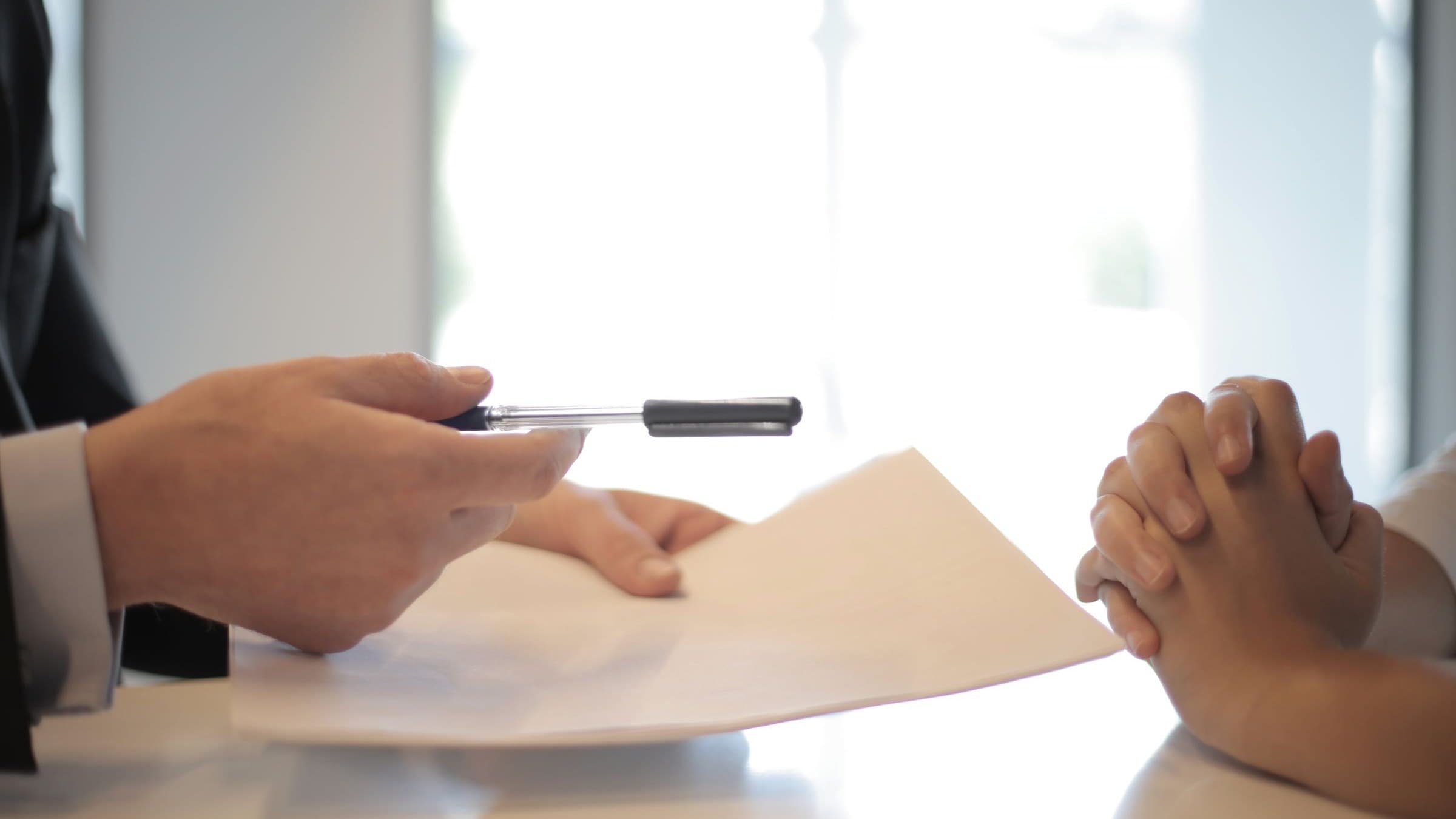 Close-up of a hand in a suit offering a pen over a document to a person with clasped hands