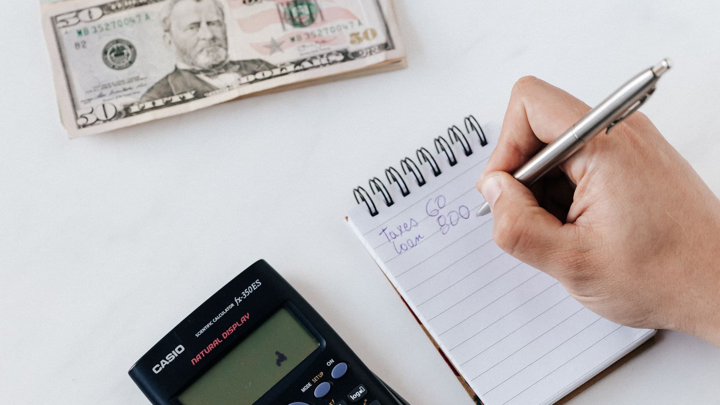 Hand writing expenses in a small notepad beside a calculator and a stack of fifty-dollar bills