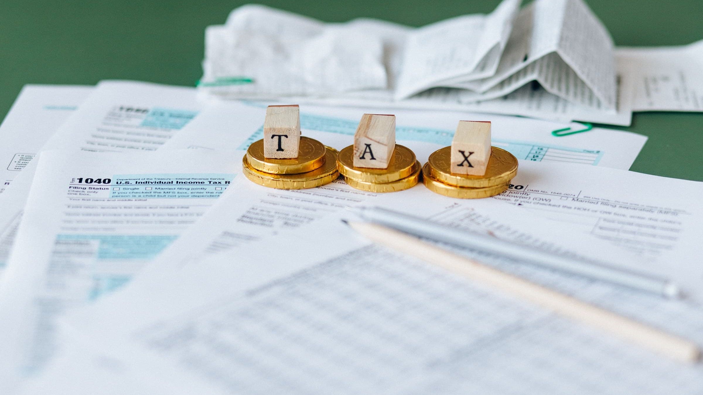 Wooden letter blocks spelling TAX on gold coins atop 1040 tax forms with a pen and crumpled receipts