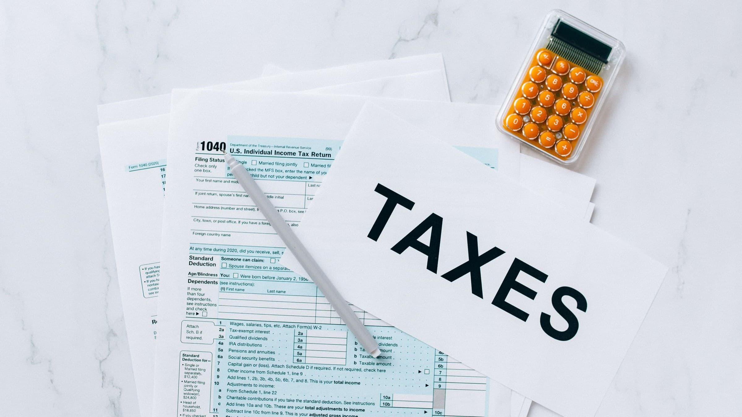 Paper labeled TAXES over a 1040 tax form with a pen and small orange calculator on marble