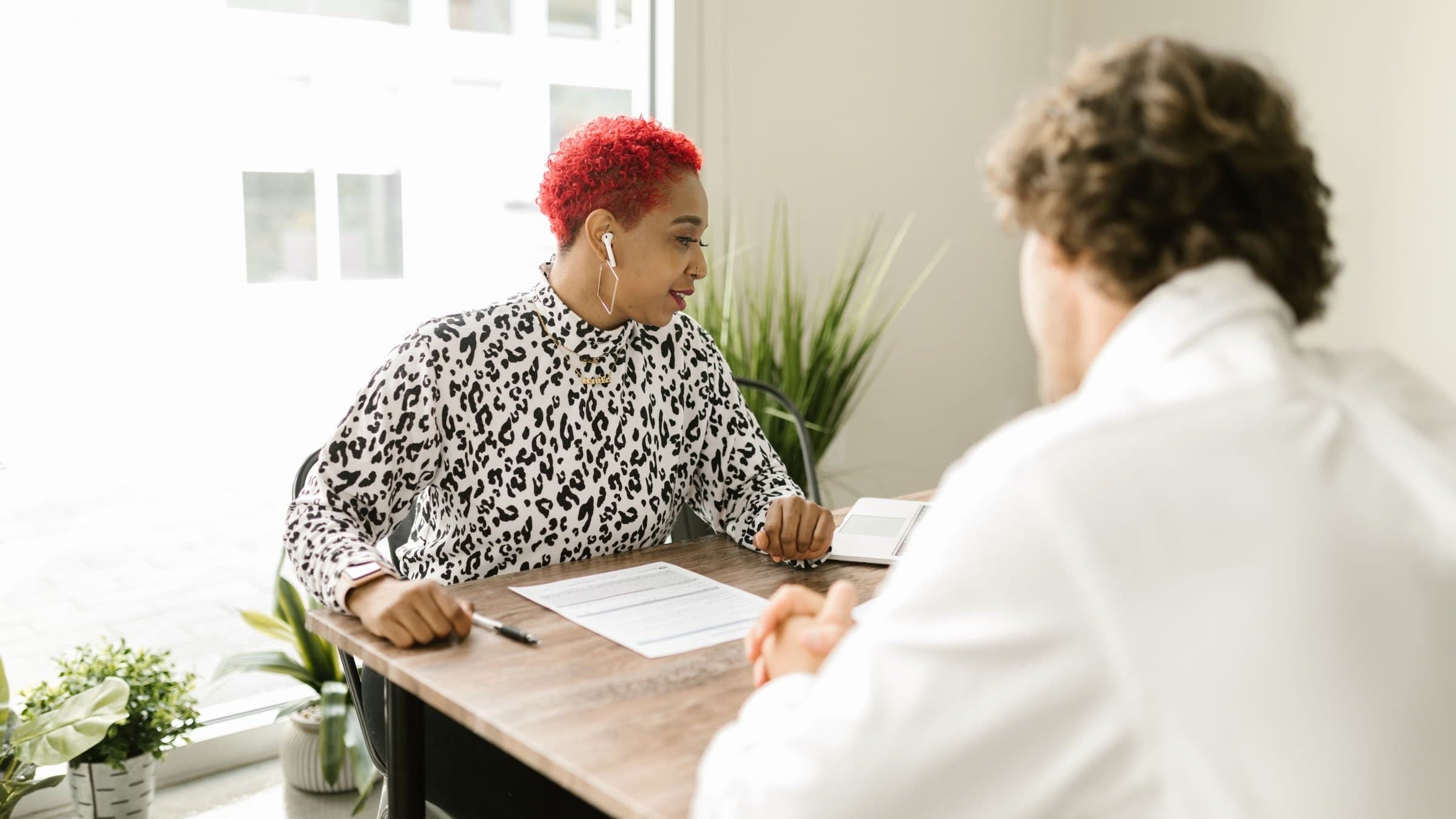 Woman with bright red hair in a leopard-print top discusses paperwork across a desk from a man