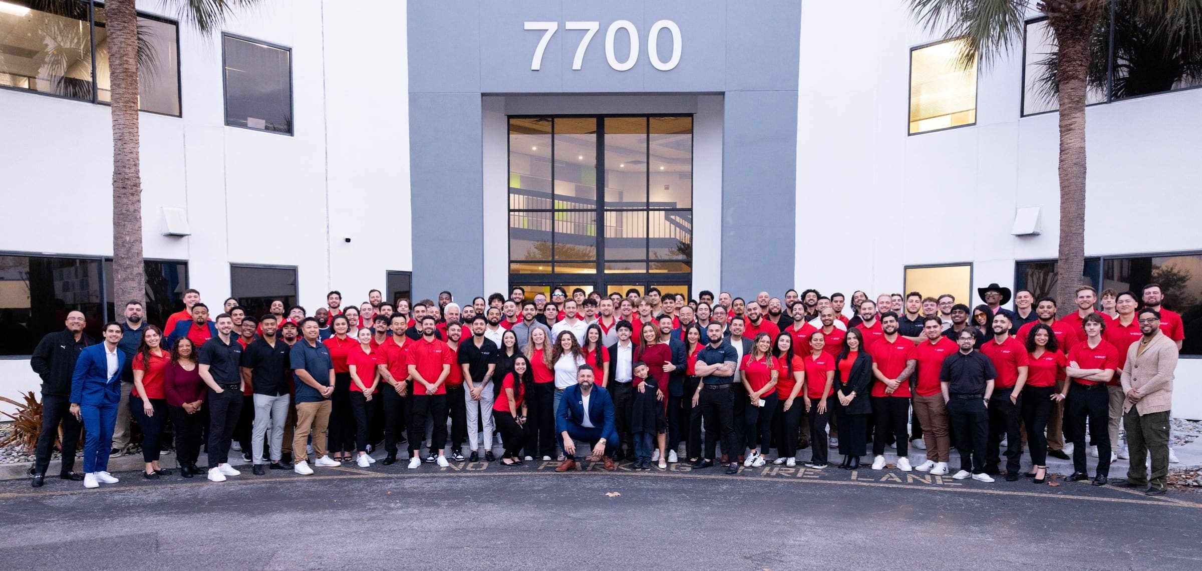 About a hundred members of the America First Healthcare team, most in red company polo shirts, standing together in front of the entrance of a white and gray office building with the number 7700 above the doors, flanked by two palm trees.